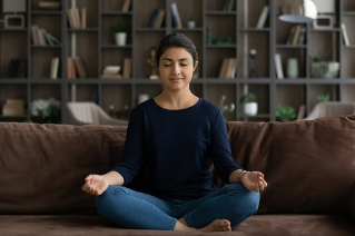 Calm millennial Indian woman sit on sofa in living room practice yoga with mudra hands. Young mixed race ethnicity female meditate relieve negative emotions relax at home. Stress free, peace concept.