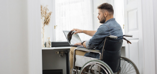 Handsome serious man in wheelchair finishing his remote work and closing modern laptop at home office. Concept of freelance, temporary disability and technology.