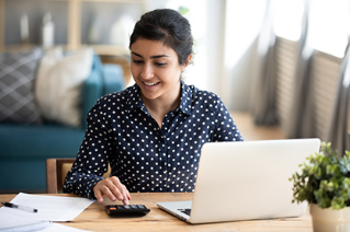 Happy millennial Indian girl sit at desk in living room manage household finances make payments on Internet on laptop, smiling young ethnic woman calculate expenditures pay bills on computer online