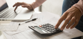 Close up cropped image young woman calculating monthly expenses, managing budget, entering data in computer application, sitting at table full of papers, loan documents, invoices, utility bills.