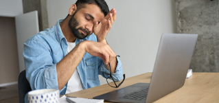 Young indian eastern tired exhausted business man rubbing eyes sitting in modern home office with laptop on desk. Overworked burnout academic Hispanic student with glasses in hand feeling eyestrain.
