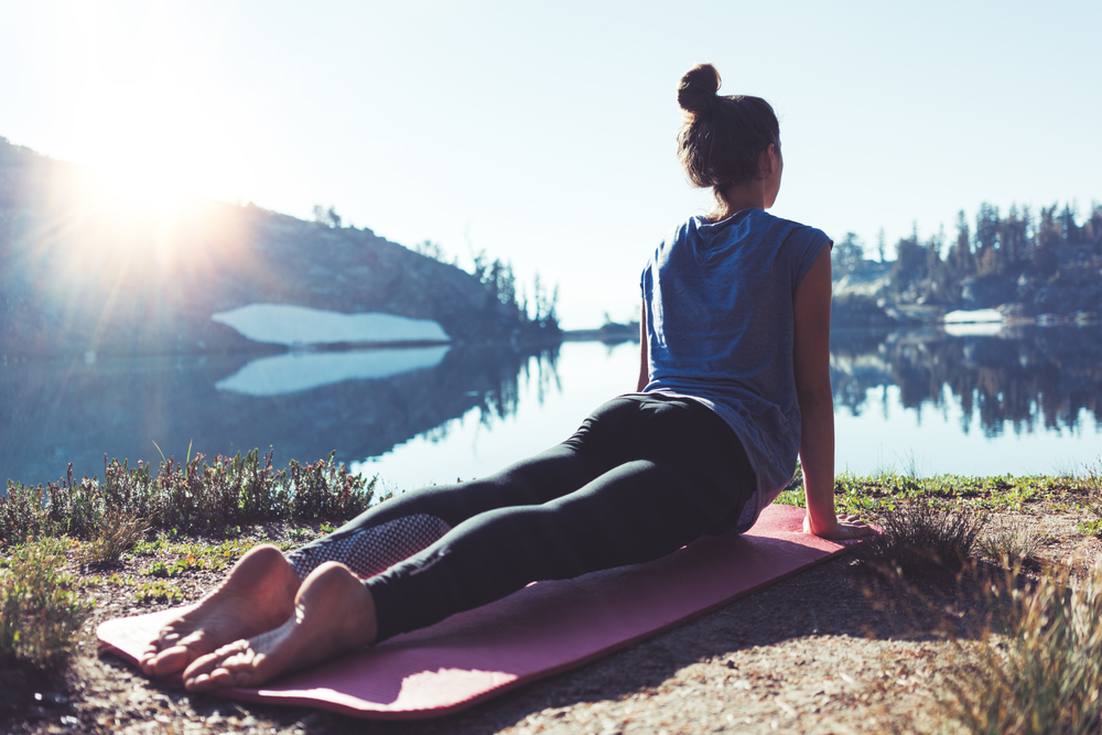 Beautiful young happy traveling girl practicing yoga outdoor in the stunning mountain wilderness in front of amazing cold lake. Calmness and relax, female happiness.